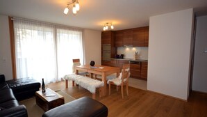 A modern living room with a wooden dining table, two chairs, a sofa, and a coffee table. There is a kitchen area with wooden cabinets and a stove. - Zell City Exclusive Lodges - TOP 4 (Zell am See)