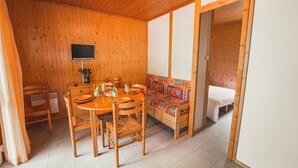 Een houten interieur met een eettafel gedekt voor vier personen, een bank met kleurrijke kussens, en een bed in de aangrenzende kamer.