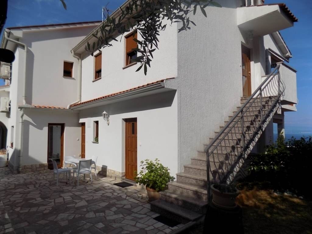 Un edificio blanco con patio de piedra, mesas al aire libre y una escalera que conduce a la entrada.