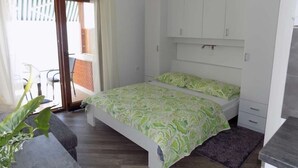 A bedroom with a bed, a small desk, a chair, a plant, and a sliding glass door leading to an outdoor area.