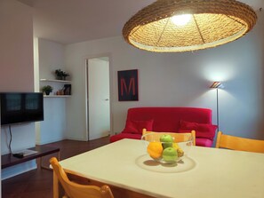A dining area with a table, chairs, a television, a red sofa, a wicker ceiling light, and a wall with a letter 'M'.