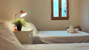Un dormitori amb un llit individual, una taula de nit amb una làmpada, una planta en test i una finestra amb persianes de fusta.