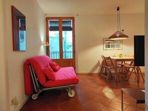 A room with a red bed, a dining table with chairs, a hanging lamp, and a window with blinds.