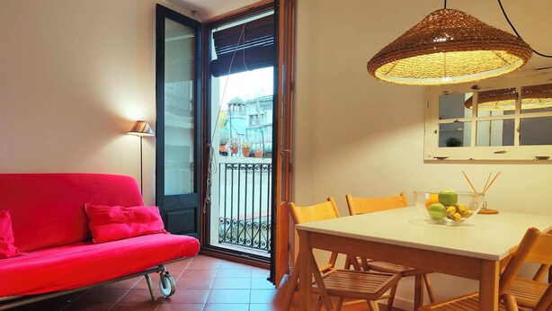 Habitació amb un sofà vermell, una taula de dinar amb cadires, un llum penjant de vímet i vista a un balcó exterior amb plantes en testos.