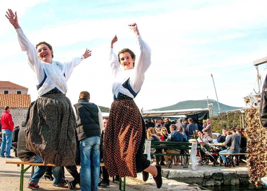 Two individuals in traditional attire dancing outdoors with a crowd in the background.