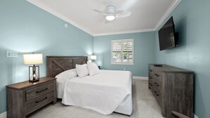 Un dormitorio con cama, mesitas de noche, cómoda, ventilador de techo, ventana con persianas y televisor montado en la pared.