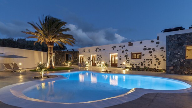 Una piscina de borde curvo, una palmera y un edificio con paredes de piedra y varias ventanas.