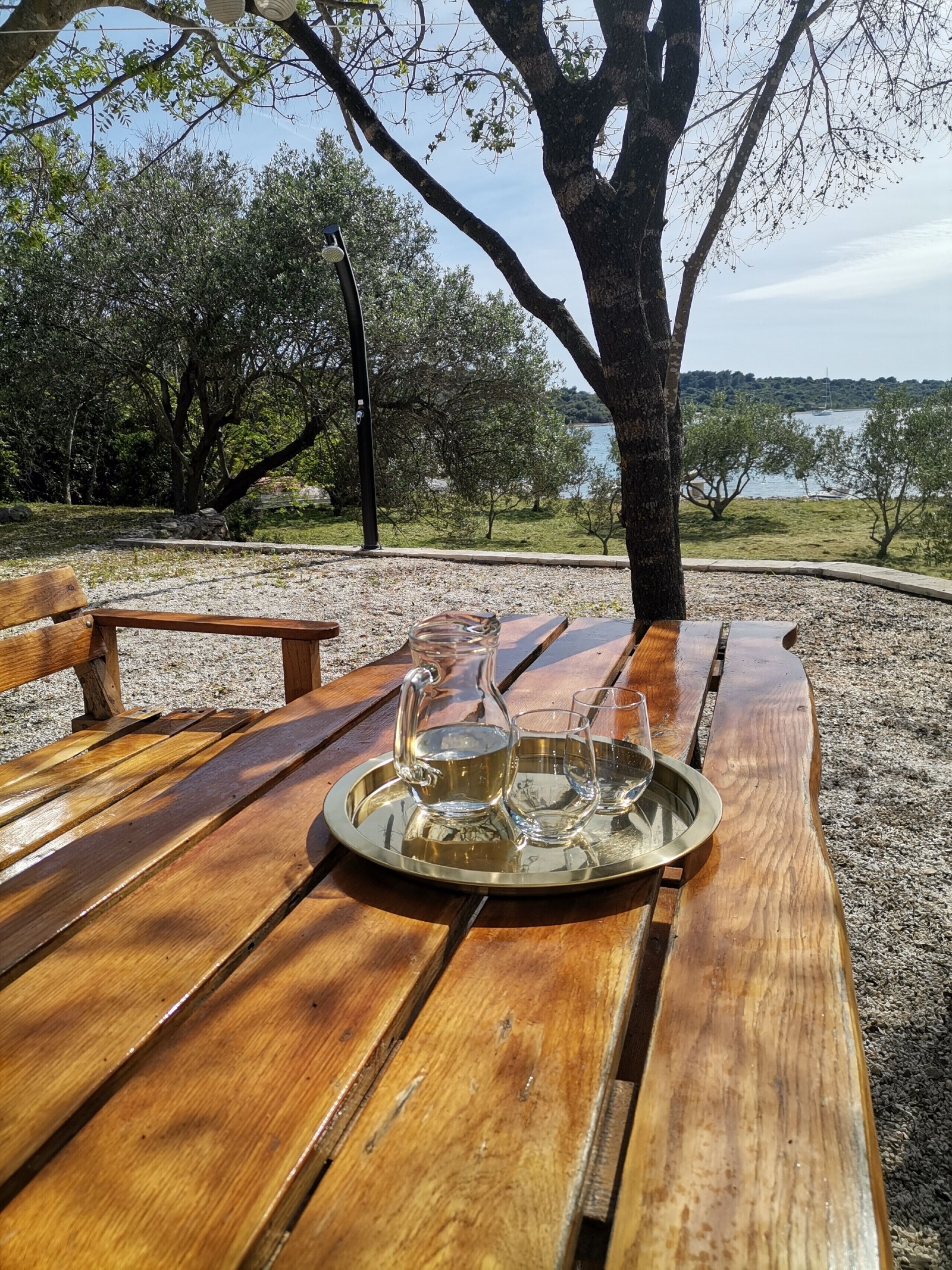 Un banc en bois sur lequel repose un plateau contenant une carafe et des verres, le tout placé devant un décor composé d’arbres et d’une étendue d’eau.