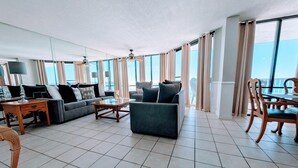 A spacious living room with a sofa, armchair, and dining table, large windows with curtains, and a ceiling fan. - Windward bldg at Edgewater, overlook the Sea Oats and Sand! (Panama City Beach)