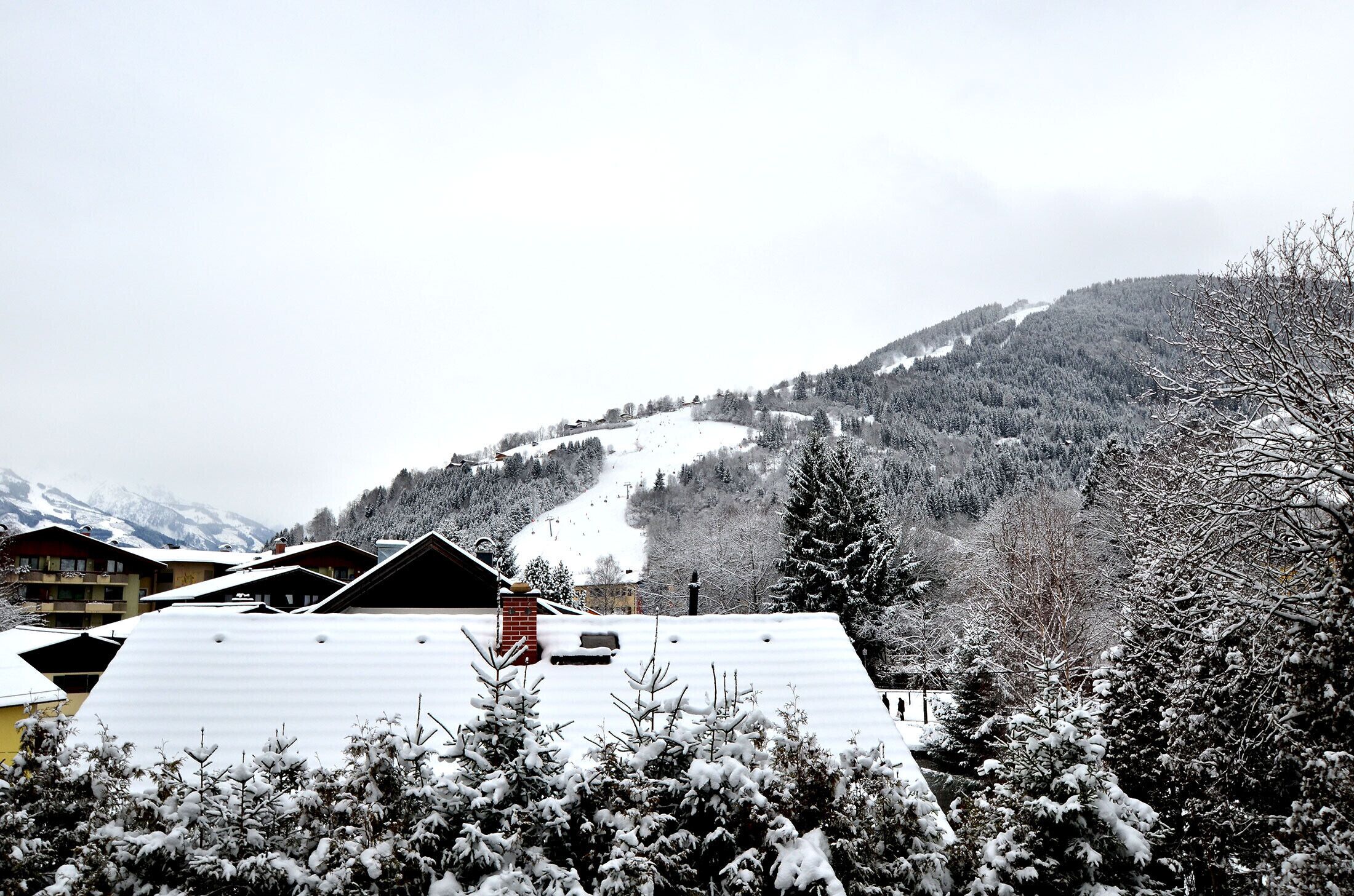A snow-covered mountain landscape with a ski slope, buildings with sloped roofs, and snow-laden trees.