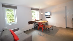 Una sala de estar moderna con un sofá gris, una mesa de comedor con sillas rojas, un televisor de pantalla plana montado en la pared y amplias ventanas con vistas a la vegetación.