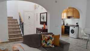 Un salon avec un canapé gris, un escalier et un coin cuisine doté de meubles en bois et d’un lave-linge.