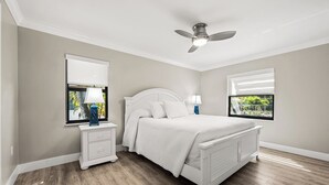 Un dormitorio con cama, mesitas de noche, ventilador de techo y dos ventanas con persianas.