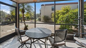 Un patio techado con una mesa y sillas, ventanas amplias y vistas a un edificio y follaje.