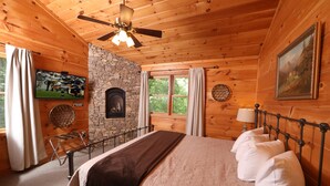 A wooden cabin bedroom with a stone fireplace, a bed with white pillows, a ceiling fan, and a television on the wall. - Shady Lady Cabin in The Smokies with Beautiful Views, Just 5 miles from Town (Sevierville)