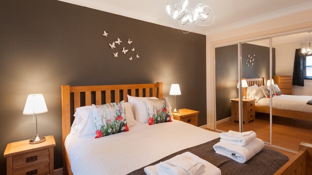 Een net opgemaakt bed met bloemenkussens, een bedlampje en een spiegel die de kamer weerspiegelt.