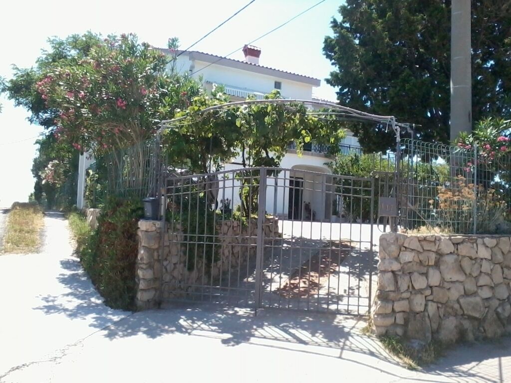 A gated entrance with a stone wall and metal gate, leading to a building with a white facade and red roof.