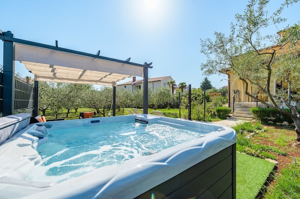 A hot tub in a backyard with a pergola, surrounded by greenery and a house in the background.