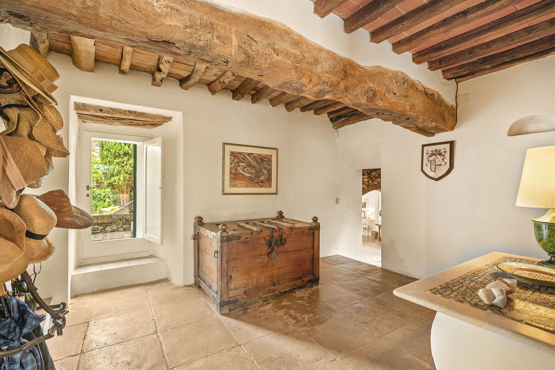 A rustic room with exposed wooden beams, a wooden chest, and a view through a door to greenery.