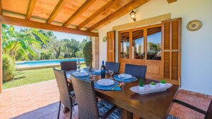 An outdoor dining area with a wooden table set for four, surrounded by wicker chairs, overlooking a swimming pool and lush greenery. - BLANCO  Ref. VP08 (Pollensa / Pollença)