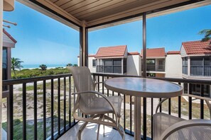 A balcony with a round table and two chairs, overlooking a beach and neighboring buildings.