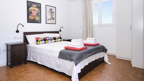 Un lit bien fait, avec du linge de lit blanc, un plaid gris et des serviettes rouges et blanches. On y trouve deux lampes fixées au mur, une table de chevet et une fenêtre avec des rideaux.