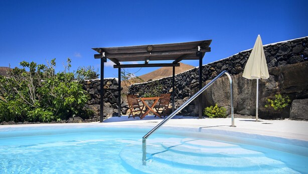Une piscine d’eau bleue et limpide, agrémentée d’une pergola en bois et d’un mur de pierre en arrière-plan.