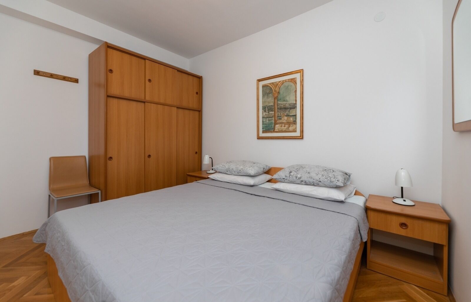 Un dormitori amb un llit, dues taules de nit, una cadira, un armari i un quadre a la paret.