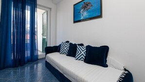 A room with a sofa, patterned pillows, a framed picture of dolphins, and a view through a window with blue curtains. - Apartment in Supetar with Terrace, Air condition, WIFI (3553-2) (Supetar)