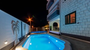 A swimming pool with a blue hue, surrounded by a white building with a brick facade and a mural of palm trees. - Apartment in Supetar with Terrace, Air condition, WIFI (3553-2) (Supetar)