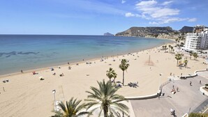 Ein Strand mit klarem, blauem Wasser, sandigem Ufer, Palmen und einer Promenade mit Gebäuden im Hintergrund.
