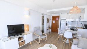 Ein modernes Wohnzimmer mit Flachbildfernseher, Glastisch und einer Couch. Es gibt einen Essbereich mit weißem Tisch und Stühlen, sowie eine Küche mit weißen Schränken und einer Pendelleuchte.