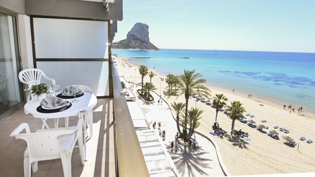 Ein Balkon mit Tisch und Stühlen mit Blick auf einen Strand, an dem Palmen und Sonnenschirme stehen.