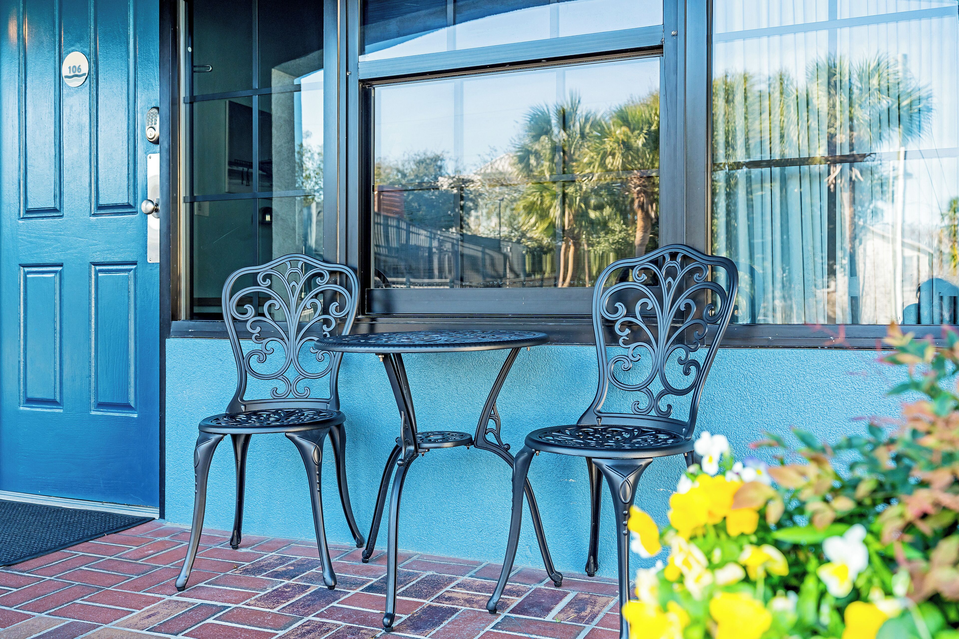 A blue door with a number plate, two metal chairs with intricate designs, a small round table, and a window with a view of palm trees and a building.