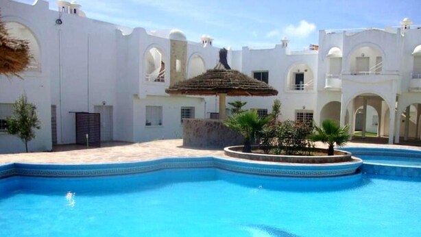 A swimming pool with a thatched roof structure, surrounded by white buildings with arches and a clear blue sky. - Nice Duplex in Sidi Bouzid 4381 (Sidi Bouzid)