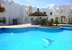 A swimming pool with a thatched roof structure, surrounded by white buildings with arches and a clear blue sky.
