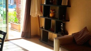 A living room with a sofa, a black shelving unit with various items, a window with a curtain, and a view of a garden outside. - Nice Duplex in Sidi Bouzid 4381 (Sidi Bouzid)