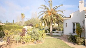 A well-maintained garden with a variety of plants and a palm tree in front of a white building with a tiled roof. - Anna - Javea, Costa Blanca (Javea)