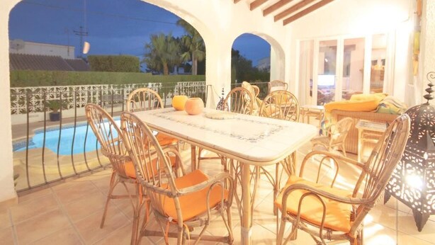 An outdoor dining area with a pool view, featuring a table and chairs, and a covered seating area with cushions. - Anna - Javea, Costa Blanca (Javea)
