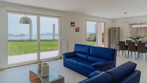 Een moderne woonkamer met een blauwe bank, een eethoek met stoelen en grote ramen met uitzicht op een brug en groen.