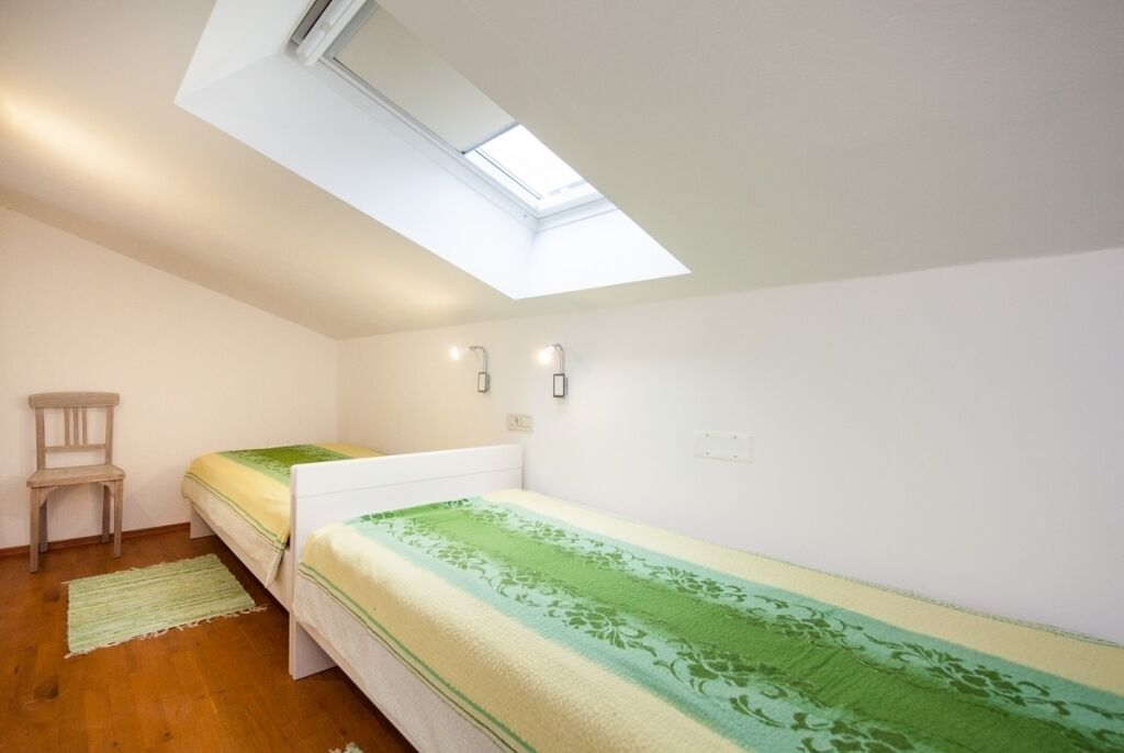 Quarto pequeno e bem iluminado, com claraboia, duas camas de solteiro, uma cadeira e um tapete pequeno.