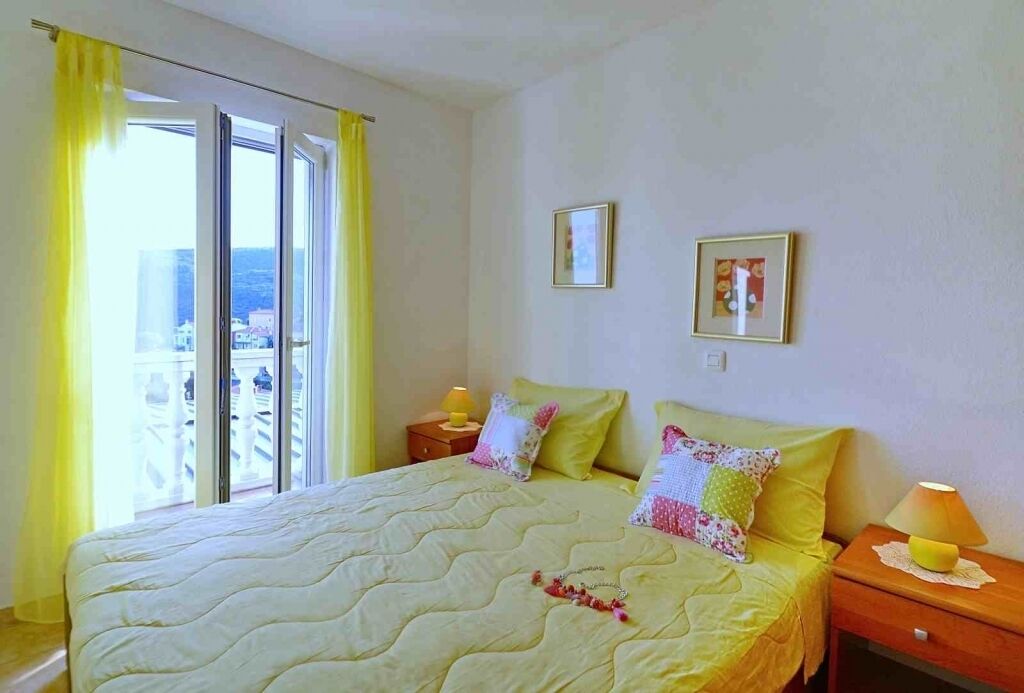A hotel room with a bed, two bedside tables, a window with yellow curtains, and a balcony view.