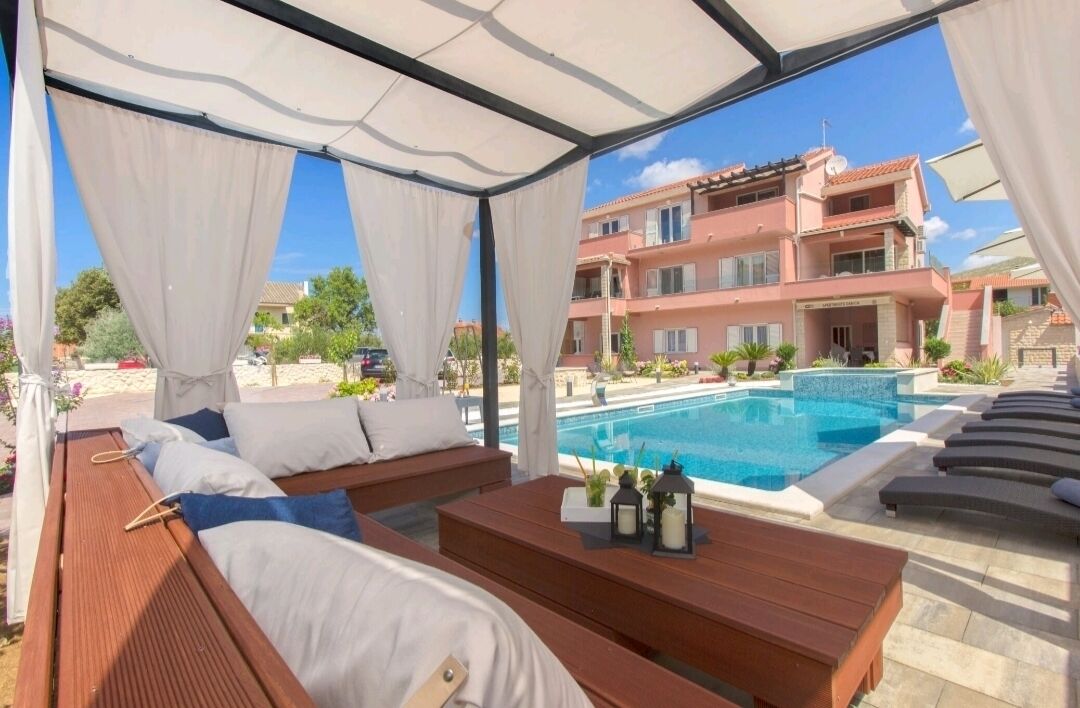 A poolside lounge area with wooden benches and a view of a swimming pool and a pink building in the background.