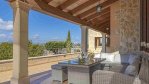A covered outdoor seating area with wicker furniture, a glass table, and a view of the landscape.
