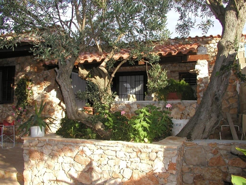 Un edificio in pietra con tetto in tegole, circondato da ulivi e piante fiorite.