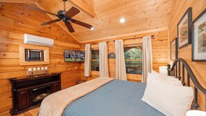 Ein Schlafzimmer in einer Holzhütte mit einem Bett, einem Deckenventilator, einer Klimaanlage, einem Kamin und einem Fenster mit Vorhängen.