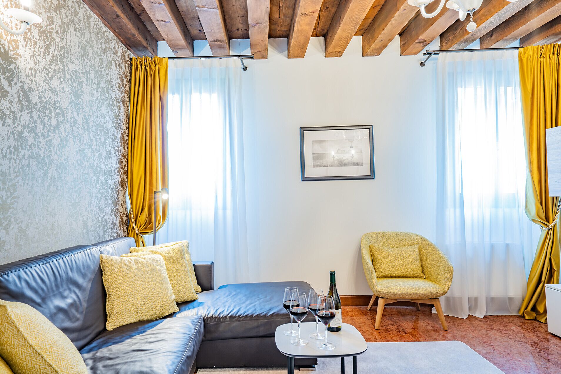 Un salon moderne avec un canapé en cuir, un fauteuil jaune, une petite table avec des verres à vin et un tableau encadré au mur.