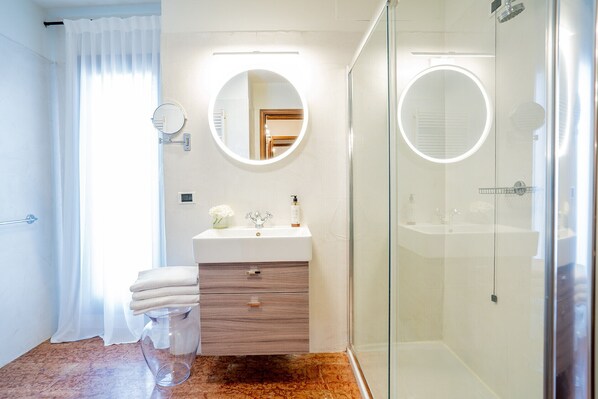 Un baño con cabina de ducha de vidrio, un lavabo blanco con tocador y un espejo redondo con luz.