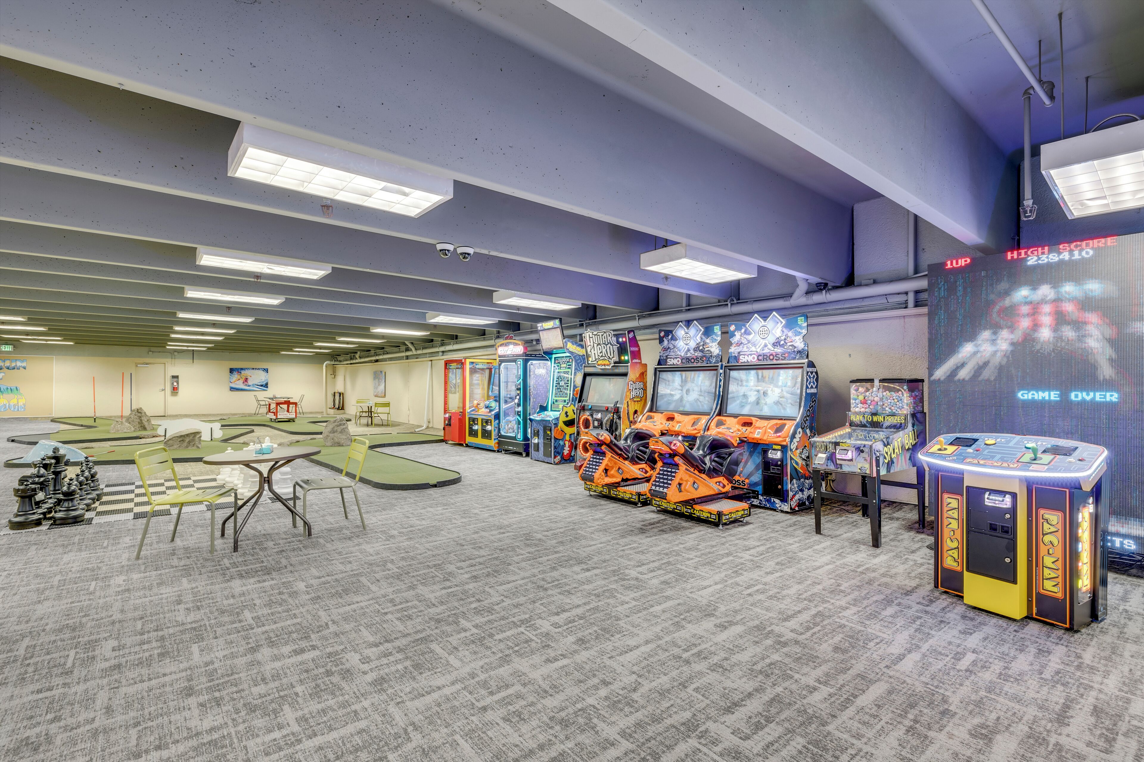 Un espace de jeux comprenant plusieurs bornes d’arcade, un jeu d’échecs et une table avec des chaises.