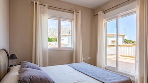 Une chambre à coucher avec un lit, deux oreillers, une table de chevet avec une lampe, et une vue sur un balcon et les bâtiments environnants.